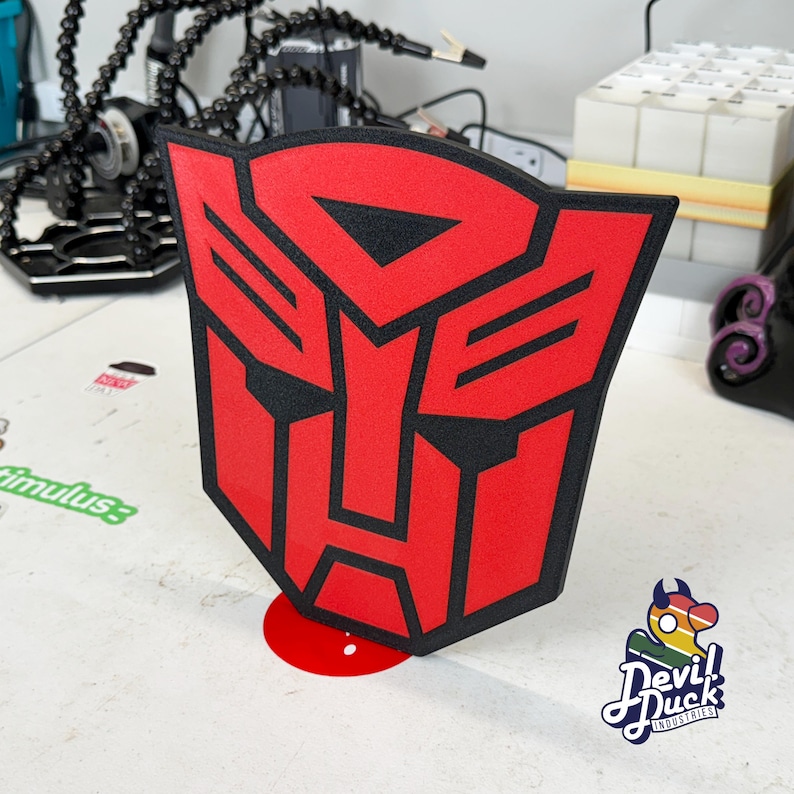 May include: A red and black 3D printed Autobot symbol, a popular symbol from the Transformers franchise. The symbol is standing on a red base.