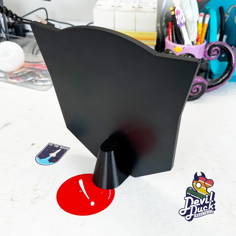 May include: Black stand with a curved top and a cone-shaped base. The stand is made of a smooth, matte material. The stand is sitting on a white surface.