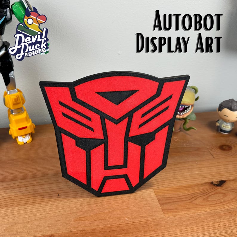 May include: Red and black Autobot logo display art. The logo is a stylized robot face with a triangular visor and a rectangular mouth.