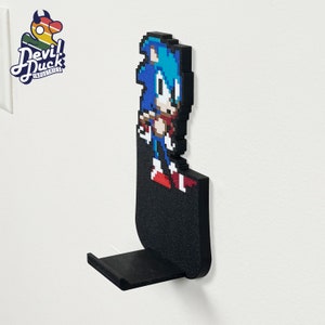 May include: A black wall-mounted phone holder featuring a pixelated image of Sonic the Hedgehog. The holder has a small shelf for a phone.