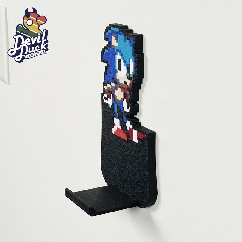 May include: A black wall-mounted phone holder featuring a pixelated image of Sonic the Hedgehog. The holder has a small shelf for a phone.