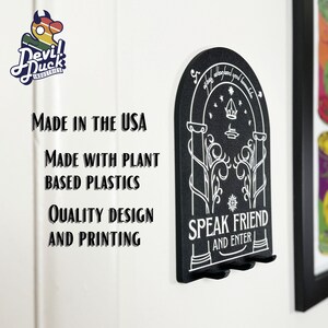 May include: Black key holder shaped like a door with the words "Speak Friend and Enter" and a design of a tree with a crown above it. The key holder is made in the USA with plant-based plastics.