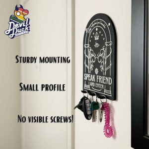 May include: Black key holder with a white design featuring a door with the text "Speak Friend and Enter". The key holder is mounted on a white wall with keys hanging from it.