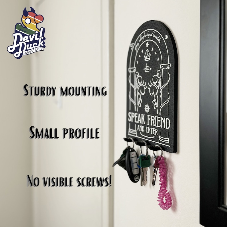 May include: Black key holder with a white design featuring a door with the text "Speak Friend and Enter". The key holder is mounted on a white wall with keys hanging from it.