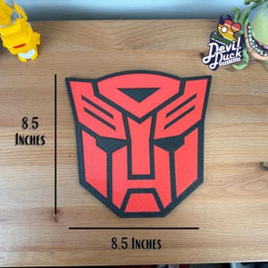 May include: A red and black Transformers logo, 8.5 inches wide and 8.5 inches tall, on a wooden surface. The logo is a stylized robot face with a black outline and red fill.