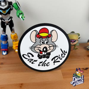 May include: A white round sticker with a black border. The sticker features a cartoon image of a gray mouse wearing a red and yellow hat. The mouse is smiling and has a cigar in its mouth. The text "Eat the Rich" is written in black around the mouse.