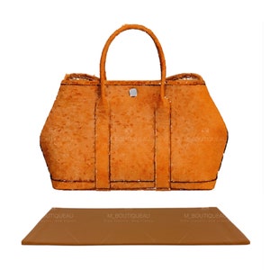 Puede incluir: Un bolso de mano naranja estructurado con superficie texturizada, dos asas superiores y un pequeño cierre metálico plateado en el centro. Debajo del bolso hay una base rectangular naranja a juego con un acabado liso y sólido.