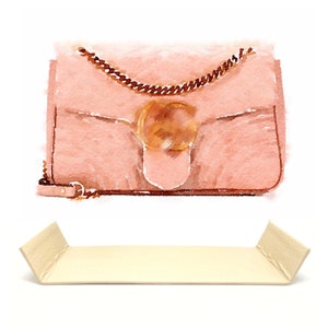 Peut inclure: Un sac à main en cuir rose clair avec une bandoulière en chaîne dorée et un grand fermoir circulaire doré. Le sac est posé sur une surface beige clair.