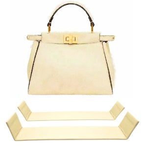 May include: A white leather handbag with a brown handle and gold hardware. Two beige fabric handbag supports are included.