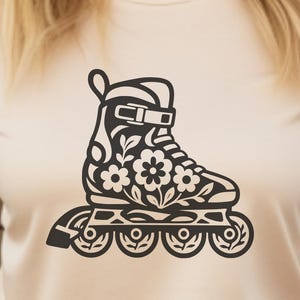 May include: A cream-colored short-sleeve t-shirt featuring a black graphic print of an inline skate decorated with floral patterns. The skate design includes visible wheels, a boot structure, and detailed flower motifs across the side panel.