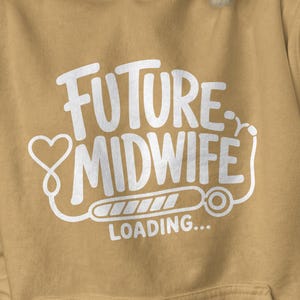 May include: A mustard yellow hooded sweatshirt featuring white screen-printed text that reads FUTURE MIDWIFE LOADING. The design includes a heart-shaped stethoscope graphic that underlines the text and connects to a loading bar icon.