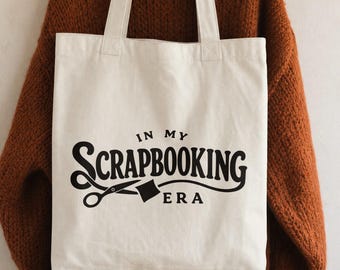 May include: A cream-colored canvas tote bag featuring black text that reads IN MY SCRAPBOOKING ERA, accompanied by a graphic of a pair of scissors. The bag has two long shoulder straps and is displayed hanging on a wooden hanger.