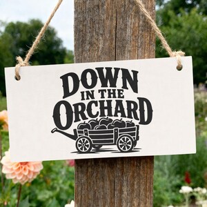 Puede incluir: Un letrero rectangular blanco colgante con texto negro que dice DOWN IN THE ORCHARD sobre un gráfico de un carro de madera lleno de manzanas. El letrero está suspendido por un cordel y tiene dos orificios circulares en las esquinas.