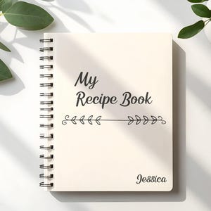 Puede incluir: Un libro de recetas con espiral de color crema con las palabras "My Recipe Book" en escritura negra. Debajo del t&iacute;tulo hay una l&iacute;nea horizontal con dise&ntilde;os de hojas. El nombre "Jessica" est&aacute; impreso en la parte inferior derecha.