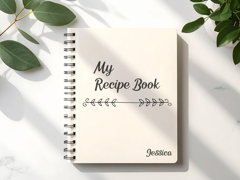 Puede incluir: Un libro de recetas con espiral de color crema con las palabras "My Recipe Book" en escritura negra. Debajo del t&iacute;tulo hay una l&iacute;nea horizontal con dise&ntilde;os de hojas. El nombre "Jessica" est&aacute; impreso en la parte inferior derecha.