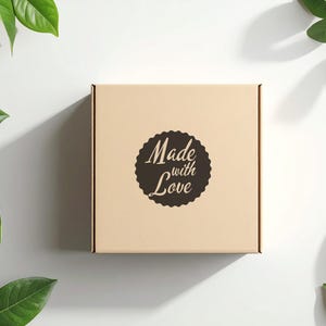 Peut inclure: Une bo&icirc;te carr&eacute;e en carton brun clair avec l'inscription "Made with Love" dans un motif circulaire marron fonc&eacute;. La bo&icirc;te est entour&eacute;e de feuilles vertes sur fond blanc.