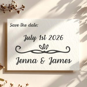 Puede incluir: Una tarjeta blanca con el texto "Save the date: July 1st 2026" en escritura negra. Debajo de la fecha hay un adorno decorativo, y los nombres "Jenna & James" est&aacute;n impresos en una escritura similar. La tarjeta est&aacute; rodeada de flores secas.