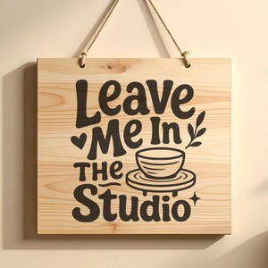 May include: A square light-coloured wooden sign hanging from a rope. The sign features dark brown text that reads Leave Me In The Studio, accompanied by a graphic of a bowl on a pottery wheel, a small heart, a leaf sprig, and a four-pointed star.
