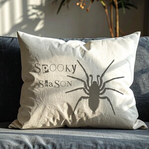 Peut inclure: Un coussin d&eacute;coratif cr&egrave;me avec les mots "Spooky Season" et une grande silhouette d'araign&eacute;e grise imprim&eacute;e. Le coussin rectangulaire repose sur un canap&eacute; gris fonc&eacute;.