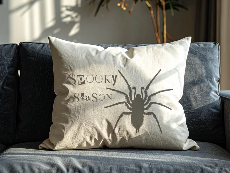 Peut inclure: Un coussin d&eacute;coratif cr&egrave;me avec les mots "Spooky Season" et une grande silhouette d'araign&eacute;e grise imprim&eacute;e. Le coussin rectangulaire repose sur un canap&eacute; gris fonc&eacute;.