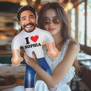 May include: An inflatable doll with a smiling face, wearing a white t-shirt with "I ❤️ SOPHIA" printed on it, and blue jeans. The doll is being held by a person wearing a white top and sunglasses.