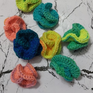 May include: A collection of nine crocheted flowers in various colors, including green, blue, yellow, orange, and pink. The flowers are arranged on a white surface with a marbled pattern.