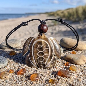 Könnte beinhalten: Eine Halskette mit einem geschnitzten Schildkrötenanhänger, schwarzem Band und Holzperle. Der Anhänger ist braun und beige mit einem Wabenmuster in der Mitte. Die Halskette wird an einem Sandstrand mit Bernsteinsteinen präsentiert.
