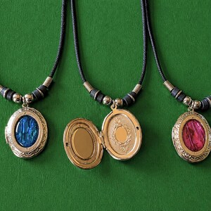 Könnte beinhalten: Drei ovale Medaillon-Halsketten mit schwarzen Kordeln und silberfarbenen Akzenten vor grünem Hintergrund. Ein Medaillon ist geöffnet und zeigt einen goldenen Innenraum. Die anderen beiden haben blaue und rosa Edelstein-Einsätze.