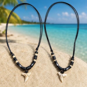 Könnte beinhalten: Zwei Haifischzahn-Halsketten mit schwarzen Kordeln und Perlen. Die Halsketten haben cremefarbene Haifischzahn-Anhänger. Präsentiert auf Sand mit tropischem Strandhintergrund.