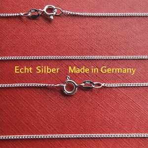 Op de afbeelding: Close-up van zilveren kettingkettingen met karabijnsluitingen tegen een rode achtergrond. De kettingen zijn fijn verbonden en lijken van sterling zilver te zijn. Tekst op de afbeelding luidt "Echt Silber Made in Germany."