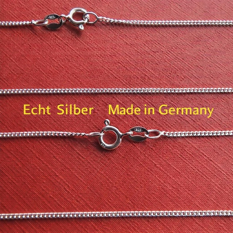 Op de afbeelding: Close-up van zilveren kettingkettingen met karabijnsluitingen tegen een rode achtergrond. De kettingen zijn fijn verbonden en lijken van sterling zilver te zijn. Tekst op de afbeelding luidt "Echt Silber Made in Germany."