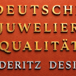 Op de afbeelding: Close-up van de woorden "DEUTSCHE JUWELIER QUALIT&Auml;T L&Uuml;DERITZ DESIGN" in gouden en zilveren letters op een getextureerde oranje achtergrond. De tekst is waarschijnlijk voor een sieradenmerk.