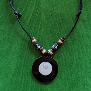 Könnte beinhalten: Eine Halskette mit einem schwarzen und weißen Muschel-Anhänger und braunen Holzperlen. Der Anhänger ist eine runde schwarze Muschel mit einer weißen spiralförmigen Muschel in der Mitte. Die Halskette besteht aus einem schwarzen Band.