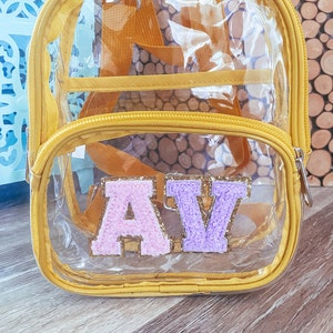 May include: A clear plastic backpack with a yellow trim. The backpack has a zipper closure and two adjustable straps. The front of the backpack has two large letters, "A" and "V", made of pink and purple glitter fabric.