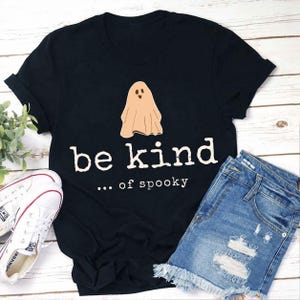 May include: A black t-shirt featuring a beige ghost illustration centered above the white text that reads be kind ... of spooky. The shirt is displayed alongside white canvas sneakers and blue denim shorts with distressed detailing.