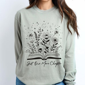May include: Sandstone-coloured long-sleeve shirt with a black floral and book graphic. The graphic features an open book with flowers and the text "Just One More Chapter". The shirt is a neutral colour.