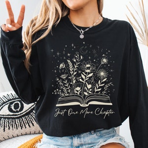 May include: Black long-sleeved sweatshirt featuring a floral and celestial design above an open book. The text "Just One More Chapter" is below the book. The word "BLACK" is at the top right.