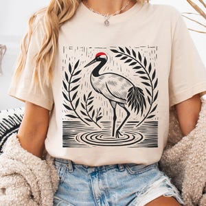 May include: A soft cream short-sleeve t-shirt featuring a black and white graphic print of a crane standing in water, surrounded by stylized leaves. The crane has a distinct red patch on its head. The text Soft Cream is printed in the top left.