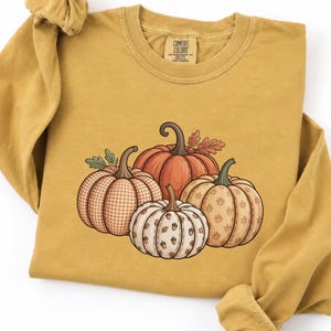 May include: A mustard yellow long-sleeve crewneck sweatshirt featuring a graphic of four pumpkins in the center. The pumpkins display various patterns including gingham, solid orange, and small leaf prints, with green stems and leaves.