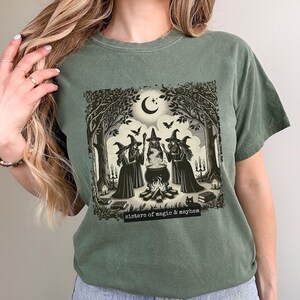 Puede incluir: Una camiseta verde con un estampado gr&aacute;fico de tres brujas de pie alrededor de un caldero en un entorno de bosque. El texto "sisters of magic & mayhem" est&aacute; impreso debajo del gr&aacute;fico.