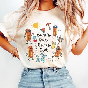 May include: A white t-shirt with a graphic design featuring hot dogs, a watermelon, a popsicle, a beach umbrella, a sun, a balloon, and the text "Sun's Out, Buns Out".