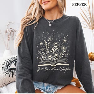 May include: A dark grey long-sleeved sweatshirt with a floral and celestial design. The graphic features an open book with flowers and stars, and the text "Just One More Chapter". The sweatshirt is a comfortable, casual style.