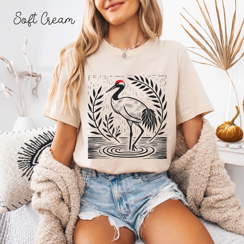 May include: A soft cream short-sleeve t-shirt featuring a black and white graphic print of a crane standing in water, surrounded by stylized leaves. The crane has a distinct red patch on its head. The text Soft Cream is printed in the top left.