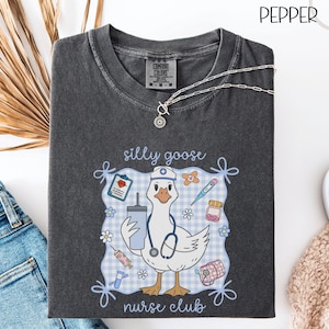 May include: Dark gray t-shirt with a graphic of a goose wearing a nurse's hat and stethoscope, holding a drink and surrounded by medical supplies. The text reads "silly goose nurse club". A silver necklace is draped on the shirt.