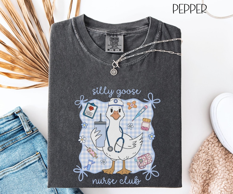 May include: Dark gray t-shirt with a graphic of a goose wearing a nurse's hat and stethoscope, holding a drink and surrounded by medical supplies. The text reads "silly goose nurse club". A silver necklace is draped on the shirt.