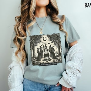 Puede incluir: Una camiseta azul claro con un estampado gr&aacute;fico de tres brujas en un entorno de bosque alrededor de un caldero. El texto "sisters of magic & mayhem" est&aacute; impreso debajo del gr&aacute;fico.
