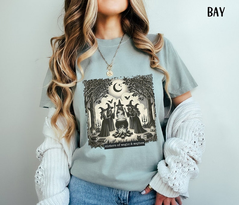 Puede incluir: Una camiseta azul claro con un estampado gr&aacute;fico de tres brujas en un entorno de bosque alrededor de un caldero. El texto "sisters of magic & mayhem" est&aacute; impreso debajo del gr&aacute;fico.