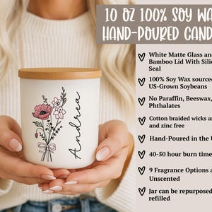 May include: A white matte glass candle with a bamboo lid, featuring a floral design and the name "Andrea." The candle is hand-poured in the USA and made with soy wax. The label reads "10 OZ 100% SOY WAX HAND-POURED CANDLE."