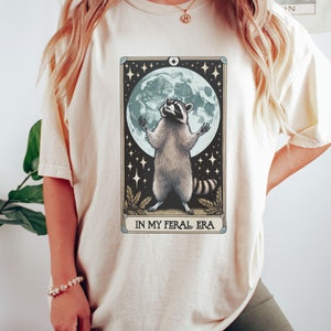 May include: A cream-colored t-shirt with a graphic of a raccoon standing on its hind legs with its paws raised in the air. The raccoon is in front of a full moon with stars in the background. The text "IN MY FERAL ERA" is printed below the raccoon.