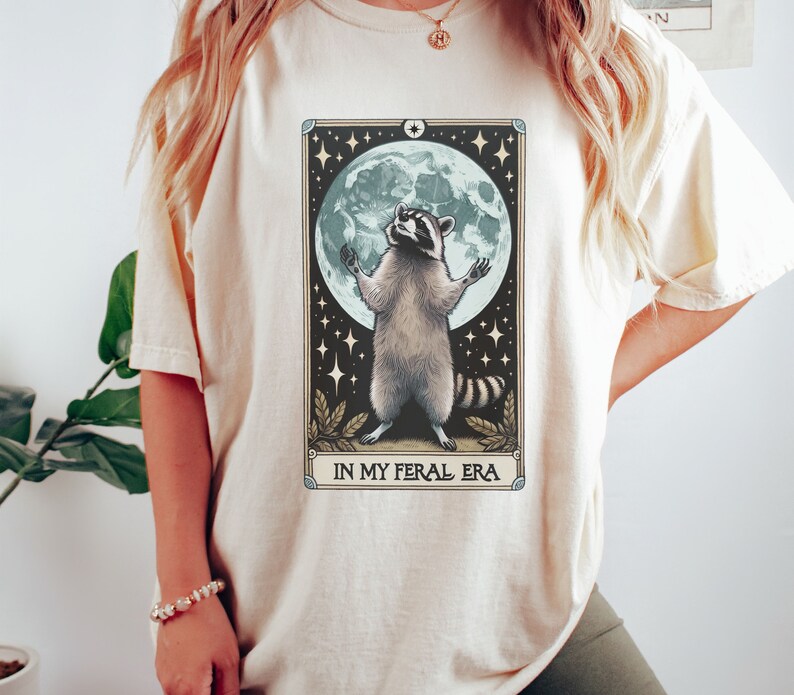 May include: A cream-colored t-shirt with a graphic of a raccoon standing on its hind legs with its paws raised in the air. The raccoon is in front of a full moon with stars in the background. The text "IN MY FERAL ERA" is printed below the raccoon.